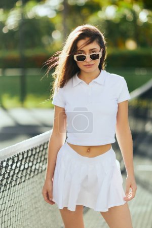 Porträt einer stilvollen jungen Frau mit brünetten langen Haaren in weißem Outfit und Sonnenbrille in der Nähe von Tennisnetz, verschwommener Hintergrund, Wind, Tennisplatz in Miami, ikonische Stadt, Florida, sonniger Tag 