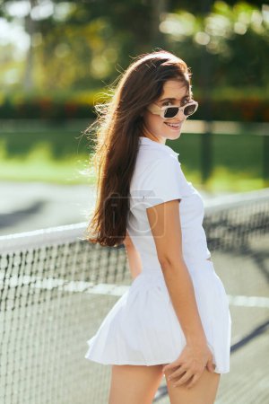 portrait de jeune femme gaie aux cheveux longs bruns debout en tenue blanche et lunettes de soleil près du filet de tennis, fond flou, vent, court de tennis à Miami, ville emblématique, Floride, journée ensoleillée 