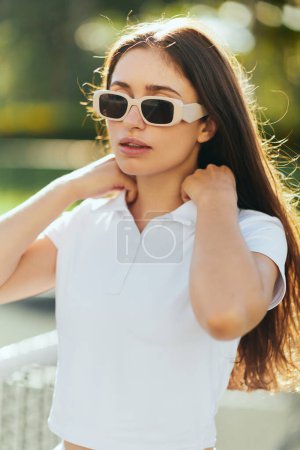 Photo for Portrait of stylish young woman with brunette long hair standing in white outfit and sunglasses near blurred tennis net on background, tennis court in Miami, iconic city, Florida, sunny day - Royalty Free Image