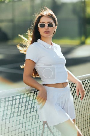 Photo for Miami, Florida, active lifestyle, beautiful young woman standing in stylish outfit and sunglasses while holding racket near tennis net, blurred background, iconic city, sunny day, vacation - Royalty Free Image