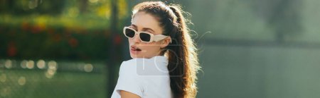 Foto de Mujer joven coqueta y seductora con cabello moreno en cola de caballo y gafas de sol de pie en polo blanco y sacando la lengua en la cancha de tenis con fondo borroso, pancarta, Miami, Florida - Imagen libre de derechos
