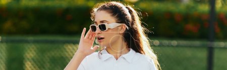 hübsche Frau mit brünetten Haaren im Pferdeschwanz trägt weißes Outfit mit Poloshirt und Sonnenbrille, während sie auf dem Tennisplatz mit verschwommenem Hintergrund wegschaut, Miami, Florida, Banner