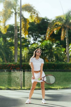 athletische Brünette mit langen Haaren im sportlichen weißen Outfit und Schläger mit Ball auf dem Tennisplatz in Miami, Florida, sonniger Tag, Palmen vor verschwommenem Hintergrund 