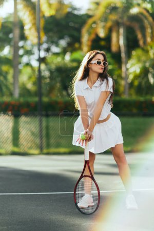 atlética morena mujer con el pelo largo posando en traje blanco deportivo y la celebración de raqueta con pelota en la cancha de tenis en Miami, Florida, día soleado, palmeras sobre fondo borroso, ciudad icónica 