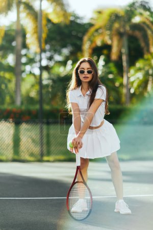 energiczna kobieta brunetka z długimi włosami stojąca w stylowym białym stroju i trzymająca rakietkę z piłką na korcie tenisowym w Miami, Floryda, słoneczny dzień, palmy na rozmytym tle, spódnica tenisowa  
