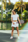 energetic woman with long hair standing in stylish white outfit and holding racket with ball on tennis court in Miami, Florida, Sunny day, palm trees on blurred background, competition  Longsleeve T-shirt #658652756