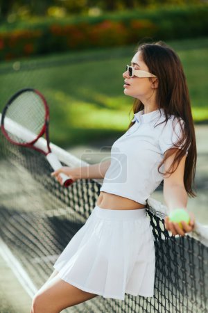 Photo for Tennis court in Miami, athletic young woman with long hair standing in white outfit and sunglasses while holding blurred racket and ball and leaning on tennis net, green background, iconic city - Royalty Free Image