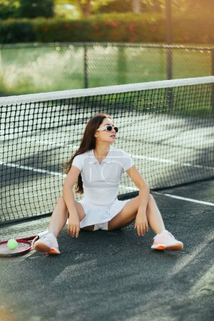 Foto de Cancha de tenis en Miami, mujer joven con estilo con pelo largo morena sentado en traje blanco y gafas de sol y descansando cerca de raqueta y pelota, red de tenis, fondo borroso, ciudad icónica, verano - Imagen libre de derechos