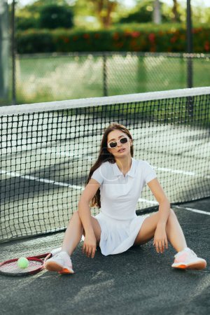 female player resting on tennis court in Miami, athletic young woman with brunette long hair sitting in white outfit and sunglasses near racket and ball, tennis net, blurred background, iconic city 