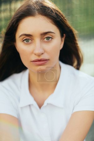 retrato de mujer joven con cabello moreno vistiendo polo blanco y mirando a la cámara después del entrenamiento en pista de tenis, red de tenis sobre fondo borroso, Miami, Florida, de cerca 