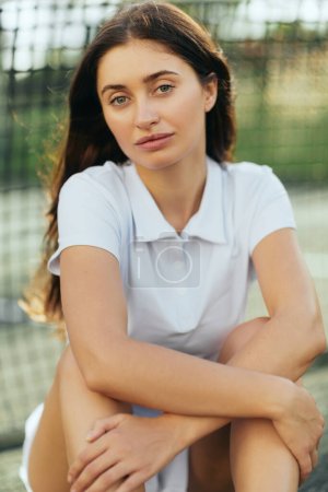 tennis court in Miami, portrait of female tennis player with brunette long hair wearing white polo shirt and looking at camera after training, tennis net on blurred background, Florida