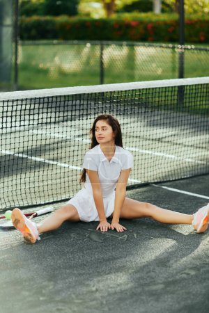 Tennisspielerin dehnt sich vor dem Spiel, junge Frau mit langen Haaren sitzt im weißen Outfit neben Schläger mit Ball und Tennisnetz, verschwommener Hintergrund, Miami, ikonische Stadt, Tennisplatz, Aufwärmen