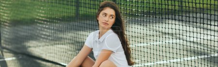 Spielerin, die in der Nähe des Tennisnetzes sitzt, junge Frau mit langen Haaren, in weißem Poloshirt, der auf dem Tennisplatz wegschaut, verschwommener Hintergrund, Miami, ikonische Stadt, körperliche Aktivität, Banner