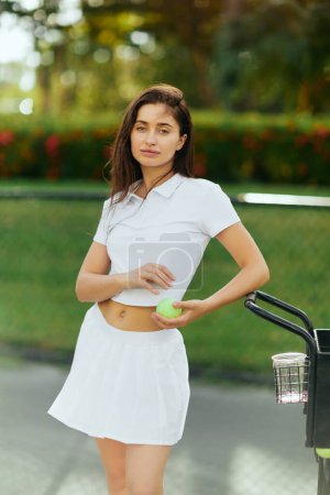 activité physique, jeune femme aux cheveux bruns debout dans une tenue élégante avec jupe et polo blanc près du chariot et tenant le ballon, fond flou, embrassé par le soleil, court de tennis à Miami 