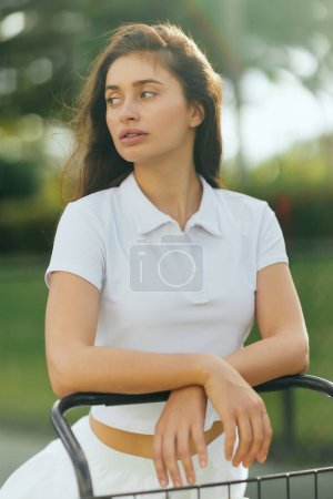 female tennis player, sporty young woman with brunette hair standing in white polo shirt near tennis cart, blurred green background, looking away, tennis court in Miami, iconic city 