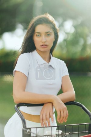 tennis court in Miami, sporty young woman with brunette hair standing in white polo shirt near tennis cart, blurred green background, looking at camera, pretty tennis player, soft filter puzzle 658653702