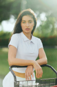 tennis court in Miami, sporty young woman with brunette hair standing in white polo shirt near tennis cart, blurred green background, looking at camera, pretty tennis player, soft filter magic mug #658653702