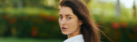 Photo for Charming and young woman with brunette long hair looking at camera on green and blurred background in Miami, vacation, summer getaway, captivating beauty, banner - Royalty Free Image