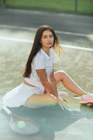female tennis player resting after game, young woman with long hair sitting in sporty outfit near racket with ball on asphalt, blurred background, Miami, tennis court, downtime, shadows, sunny day