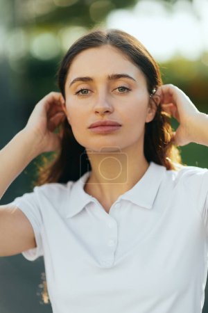 portrait of stylish young woman with brunette long hair standing in white polo shirt and looking at camera, blurred background, Miami, Florida, iconic city, natural makeup
