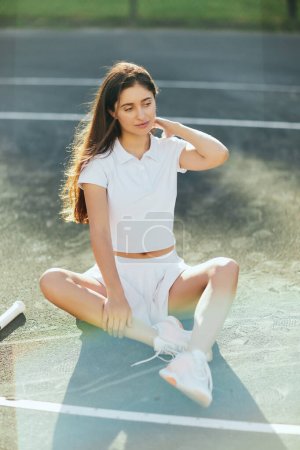 Photo for Pensive young woman with long hair sitting in white outfit near tennis racket on asphalt, blurred background, Miami, tennis court, downtime, shadows, sunny day, high angle view, looking away - Royalty Free Image