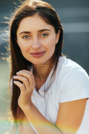 portrait of happy young woman with brunette long hair posing in white polo shirt and looking at camera, blurred background, Miami, Florida, iconic city, natural makeup 