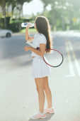 female tennis player drinking water, young woman with long brunette hair standing in white sporty outfit and holding racket on urban street in Miami, blurred background, healthy habits  Stickers #658654218