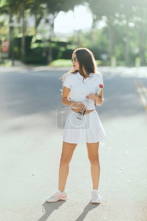Tennisspielerin mit langen brünetten Haaren in weißem Sportoutfit und Sonnenbrille mit Schläger und Wasserflasche auf urbaner Straße in Miami, verschwommener Hintergrund, gesunde Gewohnheiten, junge Frau 