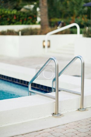 Foto de Piscina al aire libre con agua azul brillante y escalera de piscina metálica con pasamanos de acero inoxidable en el complejo hotelero de lujo, fondo borroso, vacaciones y concepto de vacaciones - Imagen libre de derechos