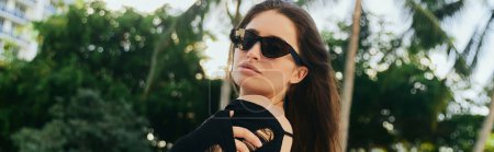 Foto de Escapada de verano, mujer morena sexy con piel bronceada, turista en vestido de punto negro y gafas de sol mirando a la cámara en el complejo de lujo durante las vacaciones en Miami, palmeras verdes, pancarta - Imagen libre de derechos