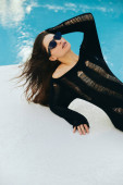 luxury resort, sexy brunette woman with tanned skin in black knitted dress and sunglasses posing next to outdoor swimming pool with shimmering water in Miami, summer getaway, youth Stickers #658655058