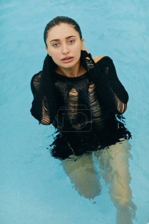 Foto de Belleza natural, sin aspecto de maquillaje, mujer morena húmeda y sexy en traje de punto negro posando dentro de la piscina al aire libre durante las vacaciones en Miami, resort de lujo, Florida - Imagen libre de derechos