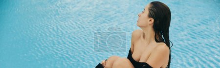 Téléchargez les photos : Détente au bord de la piscine, brunette femme sexy en robe noire avec les épaules nues assis à côté de la piscine extérieure avec de l'eau chatoyante à Miami, sensualité, mode de villégiature, embrassé au soleil, bannière - en image libre de droit