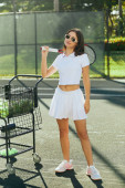 pretty tennis player in sunglasses, young woman with brunette hair standing in white outfit with skirt and polo shirt near cart with balls, blurred background, sun-kissed, tennis court in Miami  magic mug #658657284