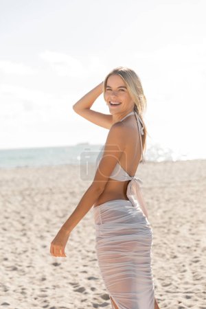 A young, blonde woman stands gracefully on a sandy Miami beach, taking in the beauty of the horizon and the vast ocean.