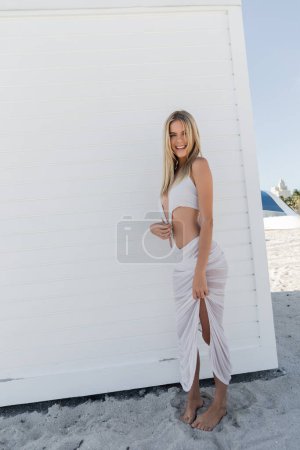 A young, beautiful blonde woman standing confidently in front of a plain white wall, exuding elegance and grace.