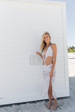 Foto de Una joven y hermosa rubia se encuentra elegantemente al lado de una pared blanca en un día soleado en Miami Beach. - Imagen libre de derechos