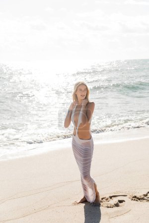 A young, beautiful blonde woman stands serenely on Miami Beach, next to the vast expanse of the ocean, embracing the peaceful moment.