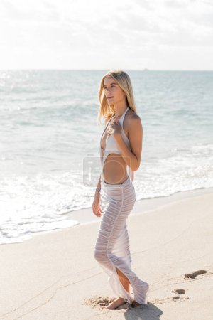 Foto de Una joven y hermosa rubia se levanta con gracia sobre una playa arenosa de Miami, empapada en la serenidad del atardecer. - Imagen libre de derechos