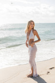 A young blonde woman stands gracefully on the sandy beach, basking in the warm Miami sun and gazing into the horizon. Stickers #665864290