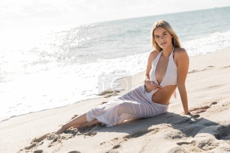A stunning blonde woman peacefully lays atop a sandy beach in Miami, embodying tranquility and relaxation.