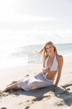 Schöne blonde Frau im weißen Bikini sitzt friedlich am Miami Beach und genießt Sonne und Sand.