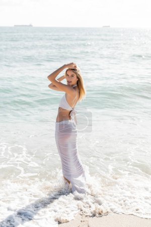 A young blonde woman standing confidently on a Miami beach, overlooking the vast ocean waves on a sunny day.