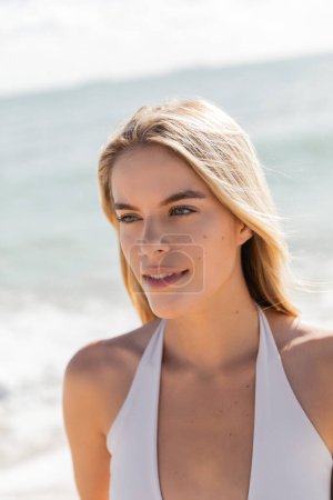 Eine junge, schöne blonde Frau im weißen Bikini steht anmutig am Strand von Miami und umarmt die ruhige Landschaft.