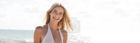 Eine junge schöne blonde Frau im weißen Bikini steht anmutig am Miami Beach und symbolisiert Frieden und Gelassenheit.