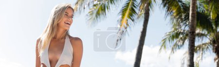 A stunning blonde woman in a white bikini stands gracefully next to a tall palm tree on a sandy Miami beach.