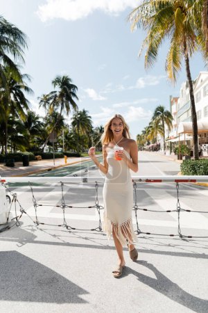 Foto de Una joven rubia con un vestido blanco que fluye delicadamente sostiene una bebida refrescante en un entorno tranquilo de Miami. - Imagen libre de derechos