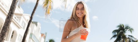 Eine junge, schöne blonde Frau in Miami hält elegant eine Tasse erfrischenden Drinks in einem weißen Kleid in der Hand.