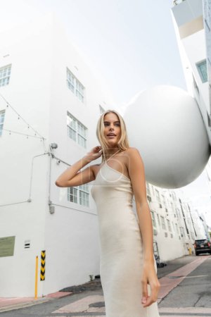 Téléchargez les photos : Une superbe femme blonde dans une robe blanche fluide prend une pose pour une photo dans un cadre vibrant de Miami. - en image libre de droit