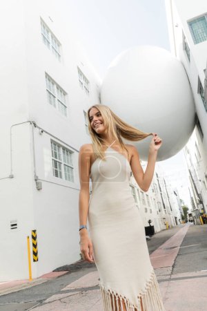A young, beautiful blonde woman in a white dress gracefully near large white balloon on a sunny day in Miami.